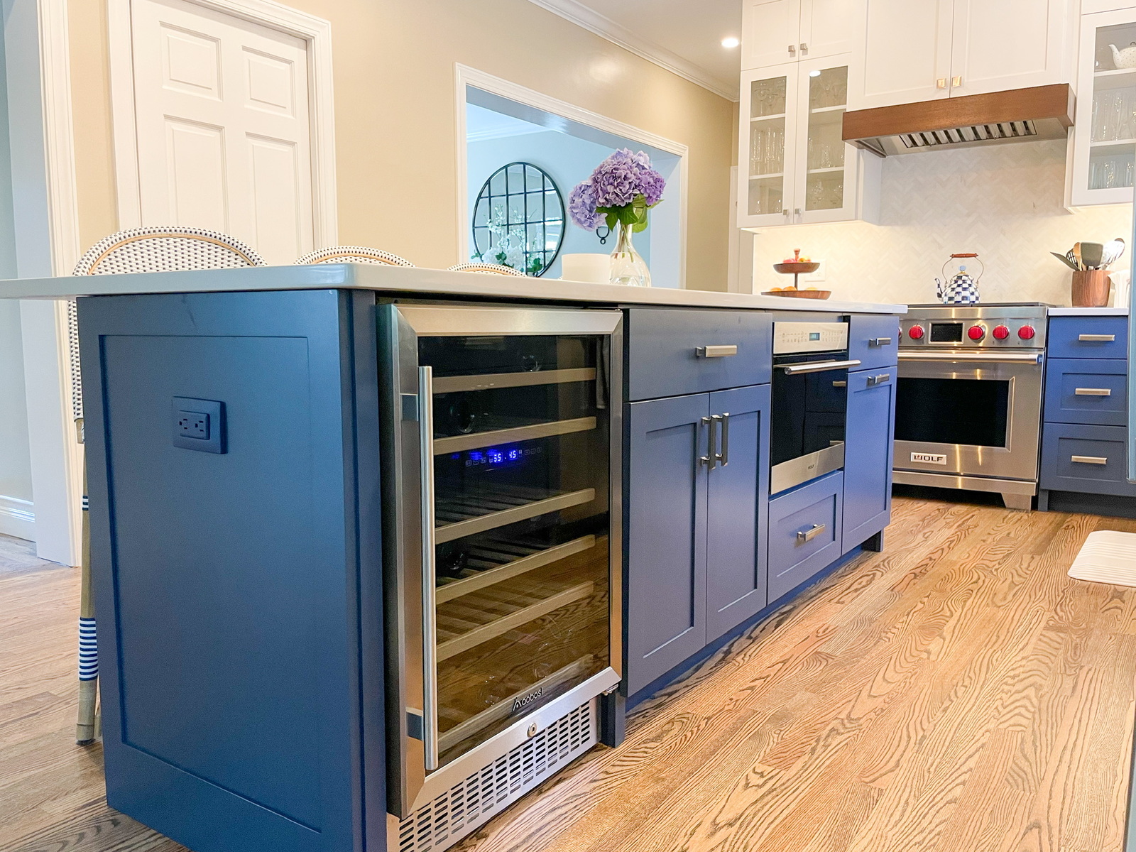 A modern kitchen featuring blue lower cabinetry and a white marble countertop. Centered in the frame is an integrated wine refrigerator filled with bottles, flanked by drawers. The background features a white subway-tile backsplash, a professional range, and light hardwood flooring.