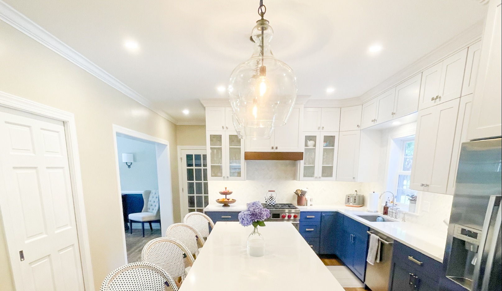 Modern kitchen with a long white marble island and gold pendant lights. Dark blue lower cabinets contrast with white upper cabinetry and a subway tile backsplash. The open space features light wood floors, a breakfast nook with blue chairs, and large windows bringing in natural light.