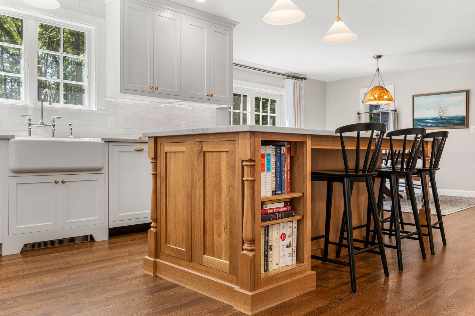A large wood kitchen island with a natural finish features built-in shelving for cookbooks and a black countertop. White perimeter cabinets with a farmhouse sink and subway tile backsplash contrast the warm wood floors. Black barstools sit under a pendant light in the dining area beyond.
