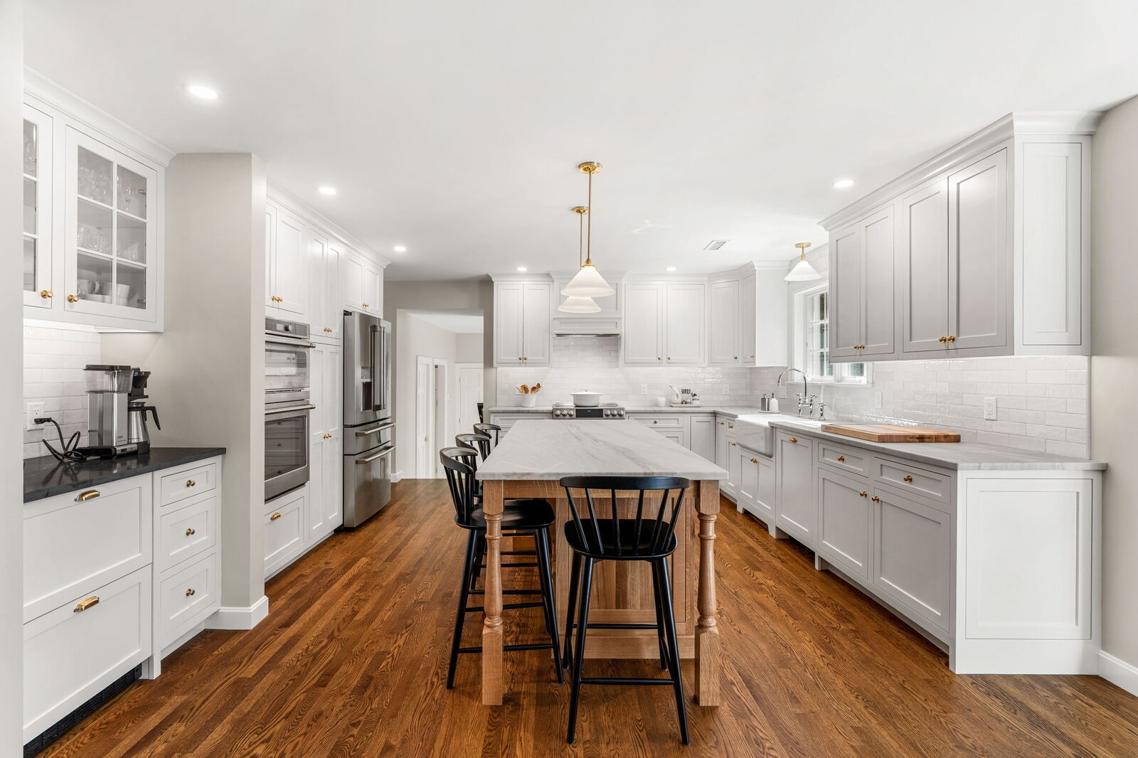 Bright, modern kitchen featuring white shaker cabinets and a large wooden central island with black stools. The space includes integrated stainless steel appliances, white marble countertops, and warm hardwood floors, creating an open and airy feel with recessed lighting.