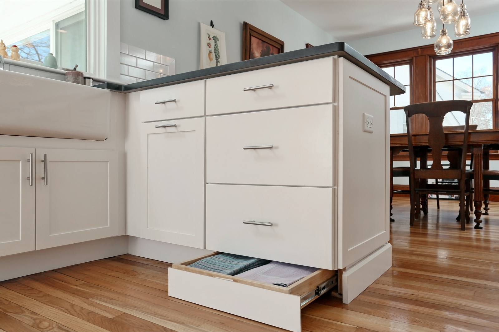 A white kitchen island with integrated toe-kick storage drawers at the base, one of which is open to reveal flat items inside. The island features shaker-style cabinetry and a dark countertop, set against warm hardwood floors and a glimpse of a dining area in the background.