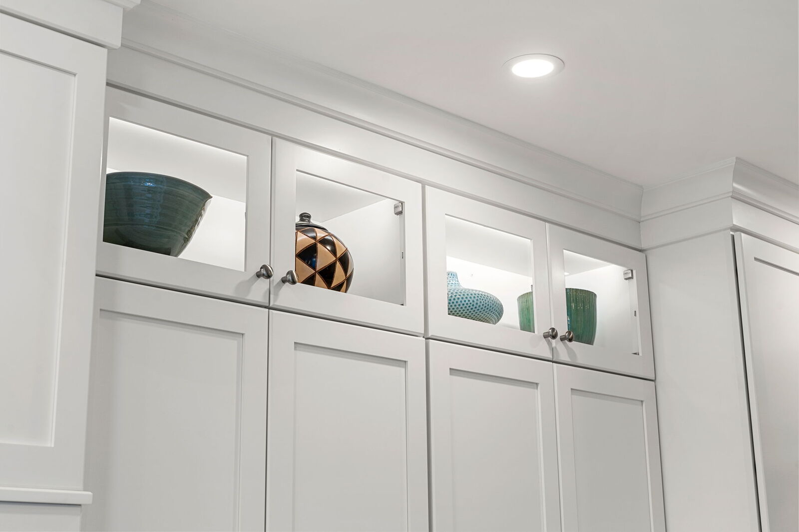 Elegant white built-in cabinetry featuring glass-front upper cabinets with interior lighting that showcases decorative items. The lower section includes solid shaker-style doors and drawers, all accented by sleek dark hardware against a soft gray wall and white crown molding.