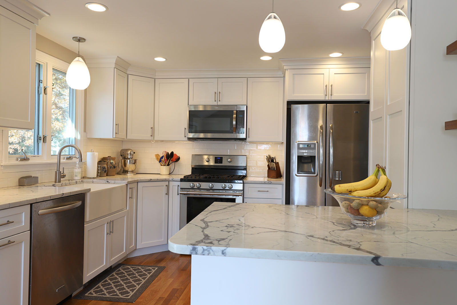 Bright, modern kitchen featuring white shaker cabinets, stainless steel appliances, and light marble countertops. A large white island with a bowl of lemons and bananas sits under glowing pendant lights. The space is completed by recessed ceiling lights and warm hardwood flooring.