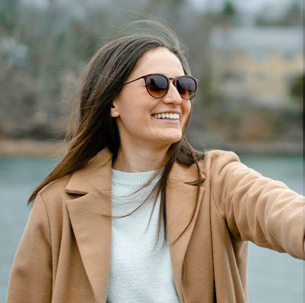 smiling-woman-in-tan-coat-and-sunglasses-enjoying-a-breezy-day-near-a-waterfront
