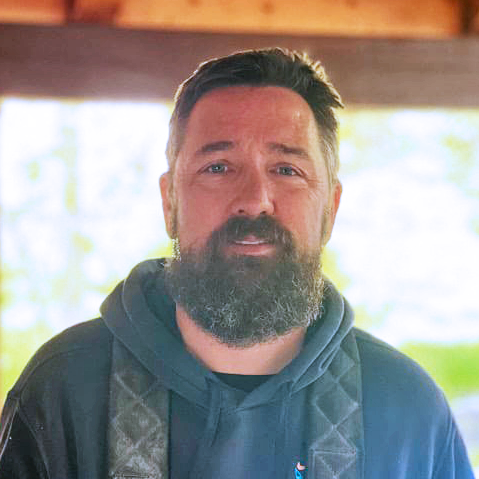 bearded-man-in-a-dark-hoodie-standing-under-a-wooden-porch-with-soft-daylight